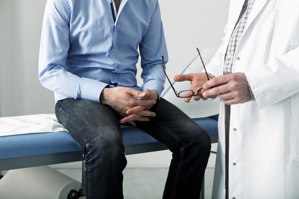 A man wearing a blue shirt and black pants sits on an exam table. A man in a white lab coat stands next to him, holding a pair of glasses.