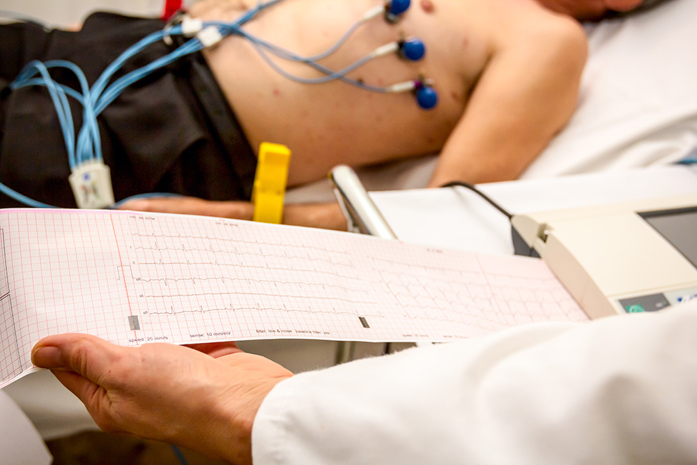 A cardiologist holds and reviews the electrocardiograph EKG diagram printed on grid paper as a patient waits in the background.