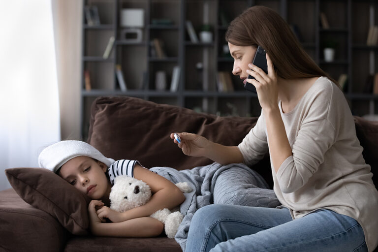 A girl lays on a couch with a washcloth on her forehead. A woman sits beside her, looking at a thermometer and talking on a cell phone.