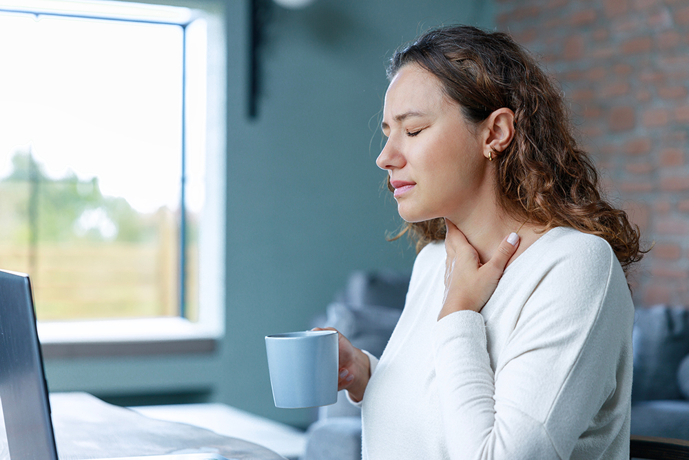 Image Alt tag: A woman in a white shirt with brown hair sits at a laptop computer. She holds a cup and is touching her throat. Her eyes are closed in apparent pain.