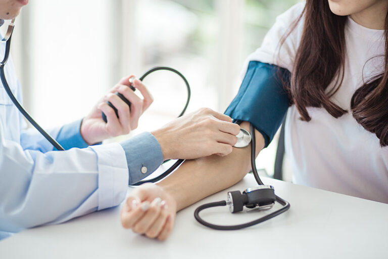 A woman sits at a table with a blood pressure cuff around her arm. A man in a white lab is inflating the cuff and listening with a stethoscope.